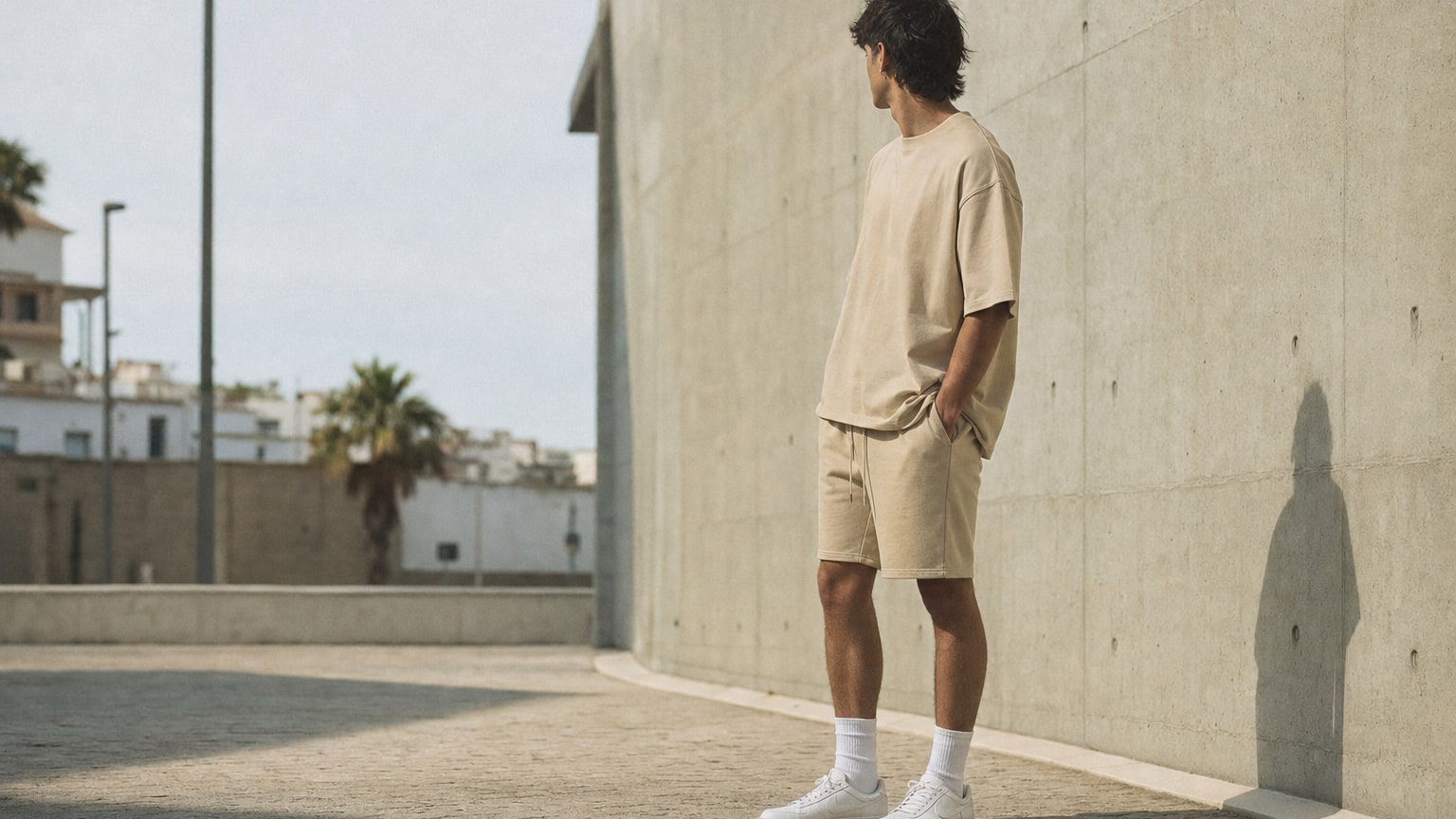 Mann in beigen Sweatshorts und oversized T-Shirt vor Betonwand, sonniger urbaner Sommer-Streetwear-Look