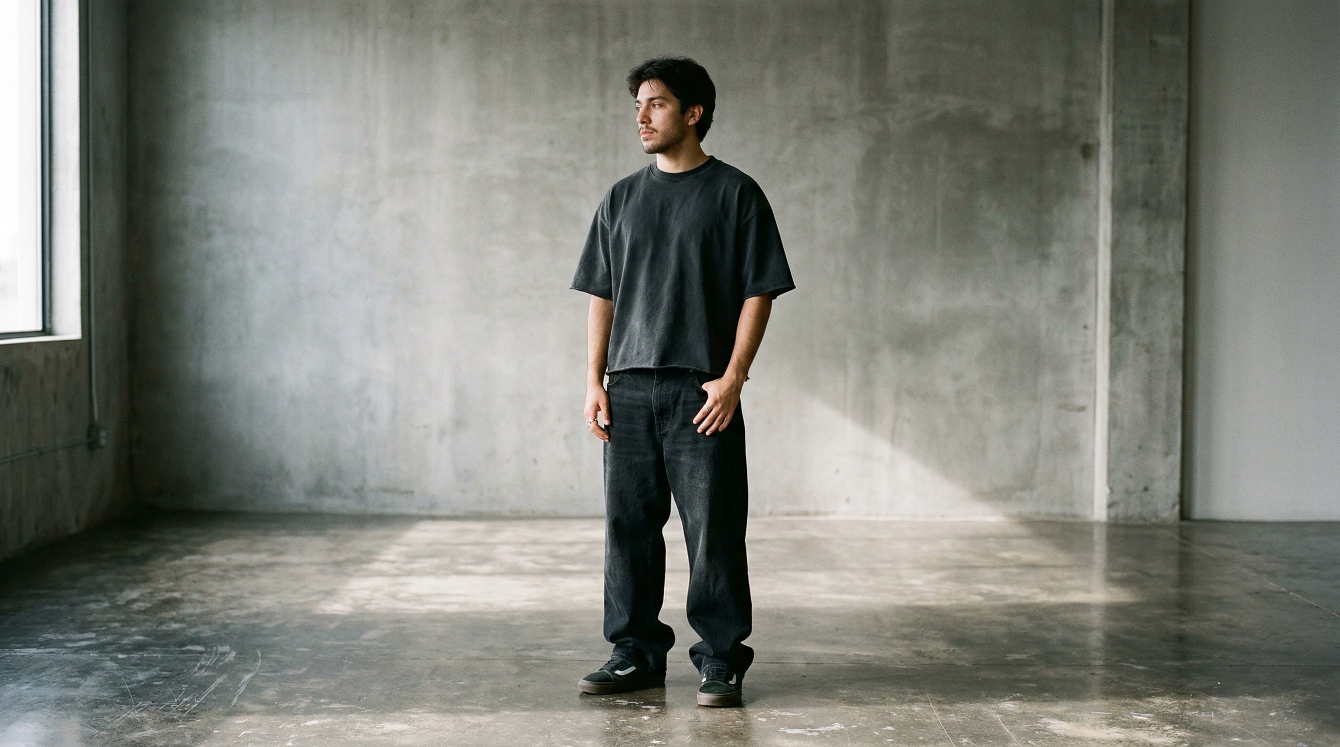 Mann in washed schwarzem Heavy Cotton T-Shirt und Baggy Denim vor grauer Betonwand, Editorial Streetwear